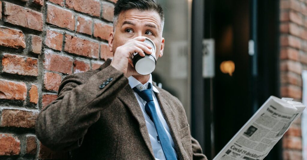 Dapper man in a formal coat drinks coffee while reading a newspaper outdoors.